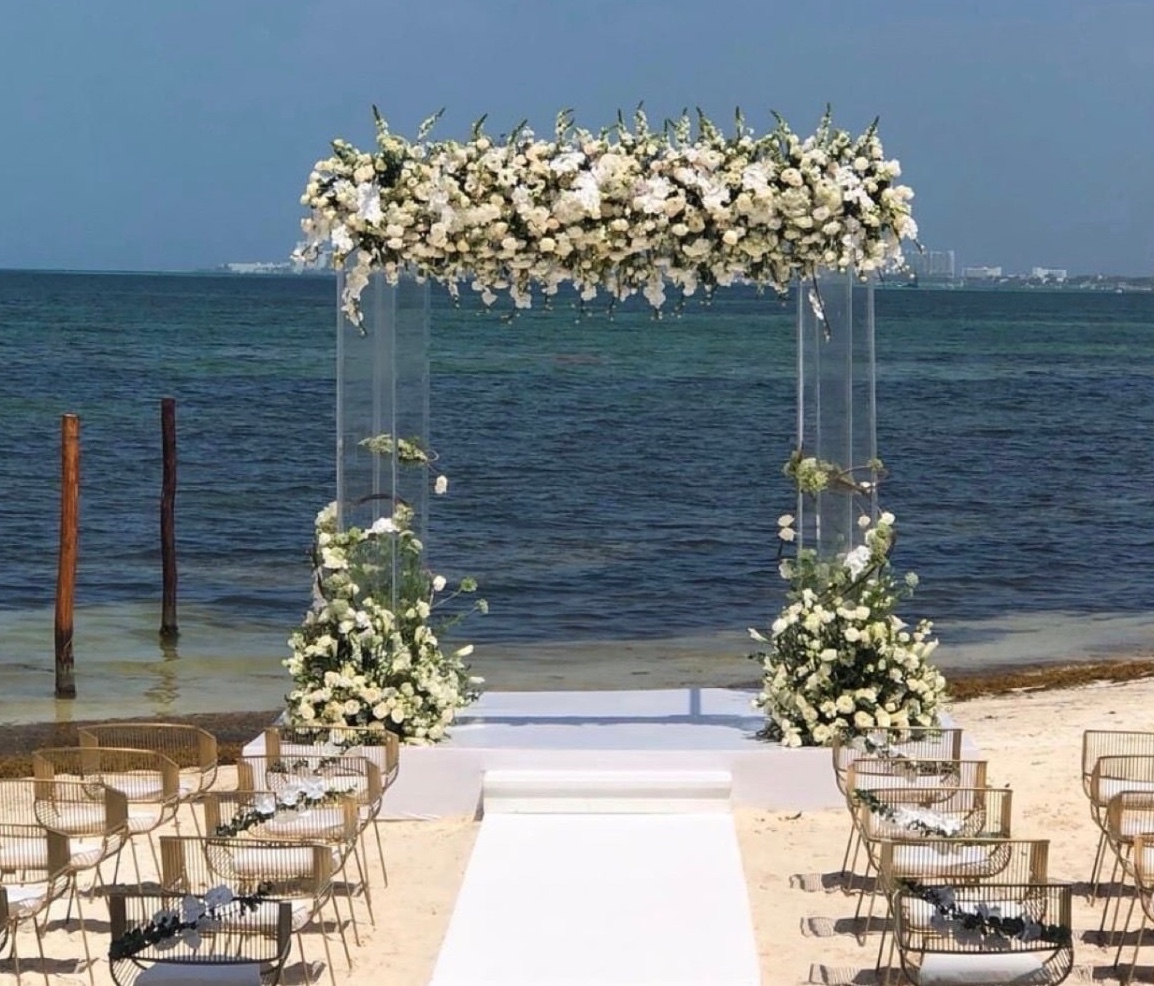 Beach ceremony with a clear arch of white florals, an aisle runner and chairs facing the water