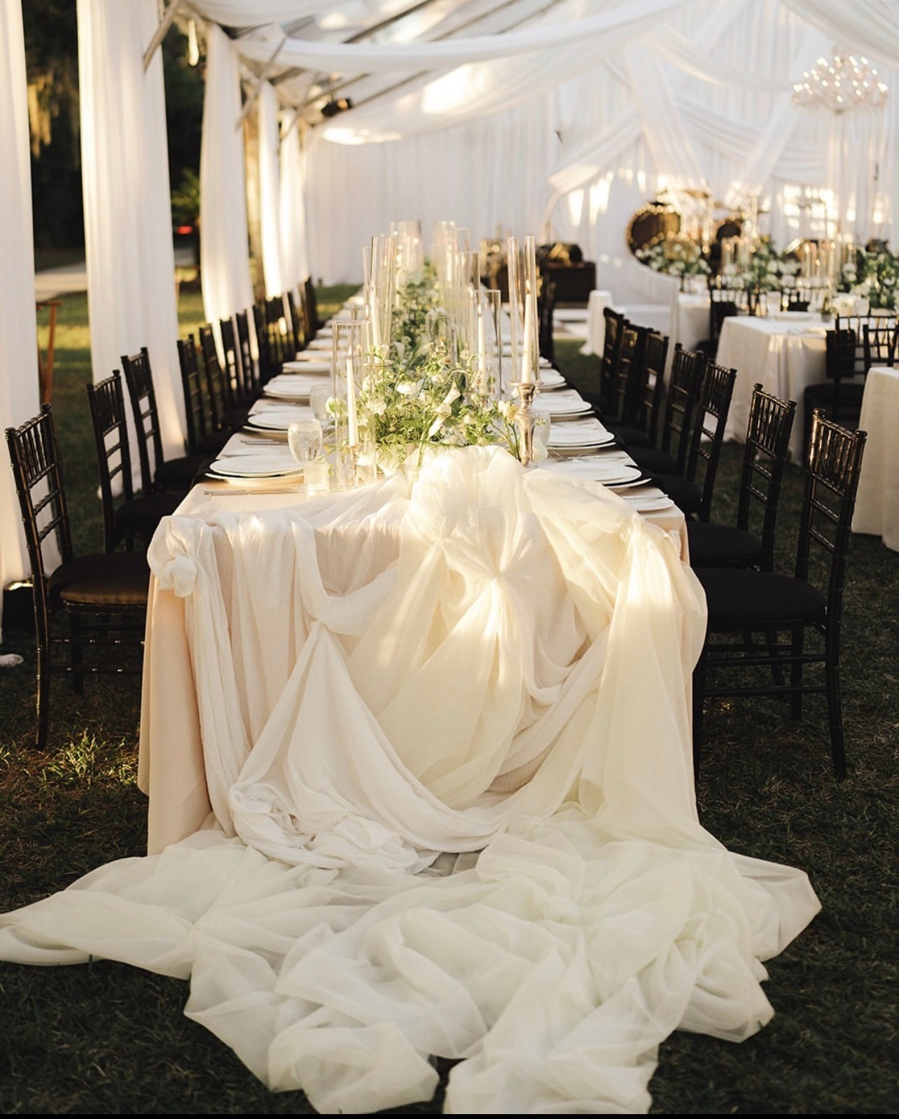 Long candlelit table with greenery under white draping, ending in a cascade of ivory fabric
